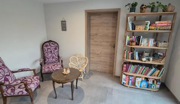 Seating area with two purple armchairs, round side table with plant, wicker shelf of books and stuffed toys, and wooden door at CHEZ GERALDINE GITES à LA FERME in Muntzenheim