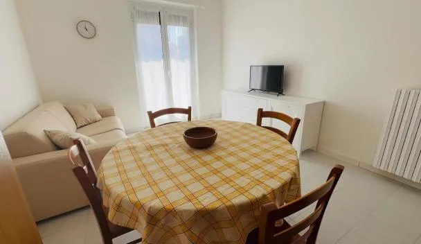 Open-plan living/dining with yellow plaid tablecloth, four wooden chairs, sofa, TV on white cabinet and balcony door at Casa Mare in Bordighera