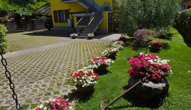 Yellow two-story house with exterior stairs, paved courtyard, lawn with pink and white petunias and an anchor planter at Appartamento L'ulivo 5 chilometri da Varenna vista lago in via privata in Lierna