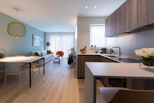A bright and contemporary open-plan hotel apartment interior combining a fully equipped kitchen, dining area, and living room in one airy space. The sleek kitchen in the foreground features dark wood-grain cabinetry, light grey stone countertops, a built-in sink with a brushed brass faucet, and a vase of fresh white tulips. A white dining table with matching chairs sits beneath a gold wire pendant light to the left. The living area beyond features a light grey sofa with olive green cushions, a terracotta accent chair, small potted plants, and a coffee table, all set against soft sage green feature walls. Light oak hardwood floors and sheer white curtains over a large window