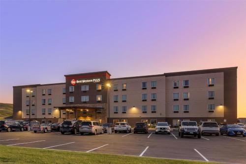 Best Western Plus Rapid City hotel exterior at dusk, with a four-story building featuring rows of windows, a stone-accented entrance, and warm outdoor lighting. The spacious parking lot in front is filled with cars, while the sky glows in soft purple and orange hues, adding a welcoming atmosphere.