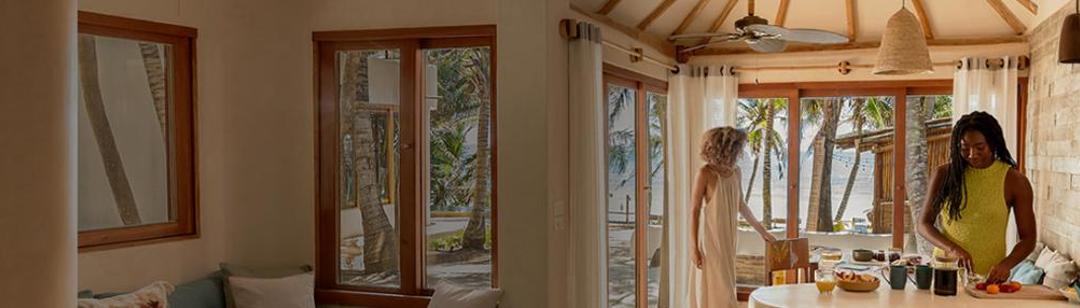 A bright, inviting kitchen with a natural wood and stone design opens up to a tropical beach view through large glass doors. Soft curtains frame the windows, letting in golden sunlight. Two women are inside. One, in a flowing dress, stands near the doors reaching toward a chair, while the other, in a summer top, chops fresh fruit at the kitchen island. On the countertop, there’s a teapot, a pineapple, and other kitchen essentials. Beyond the doors, palm trees and a sandy beach stretch toward the ocean.
