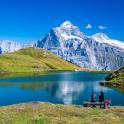 two people sitting on a picnic table near a mountain lake
