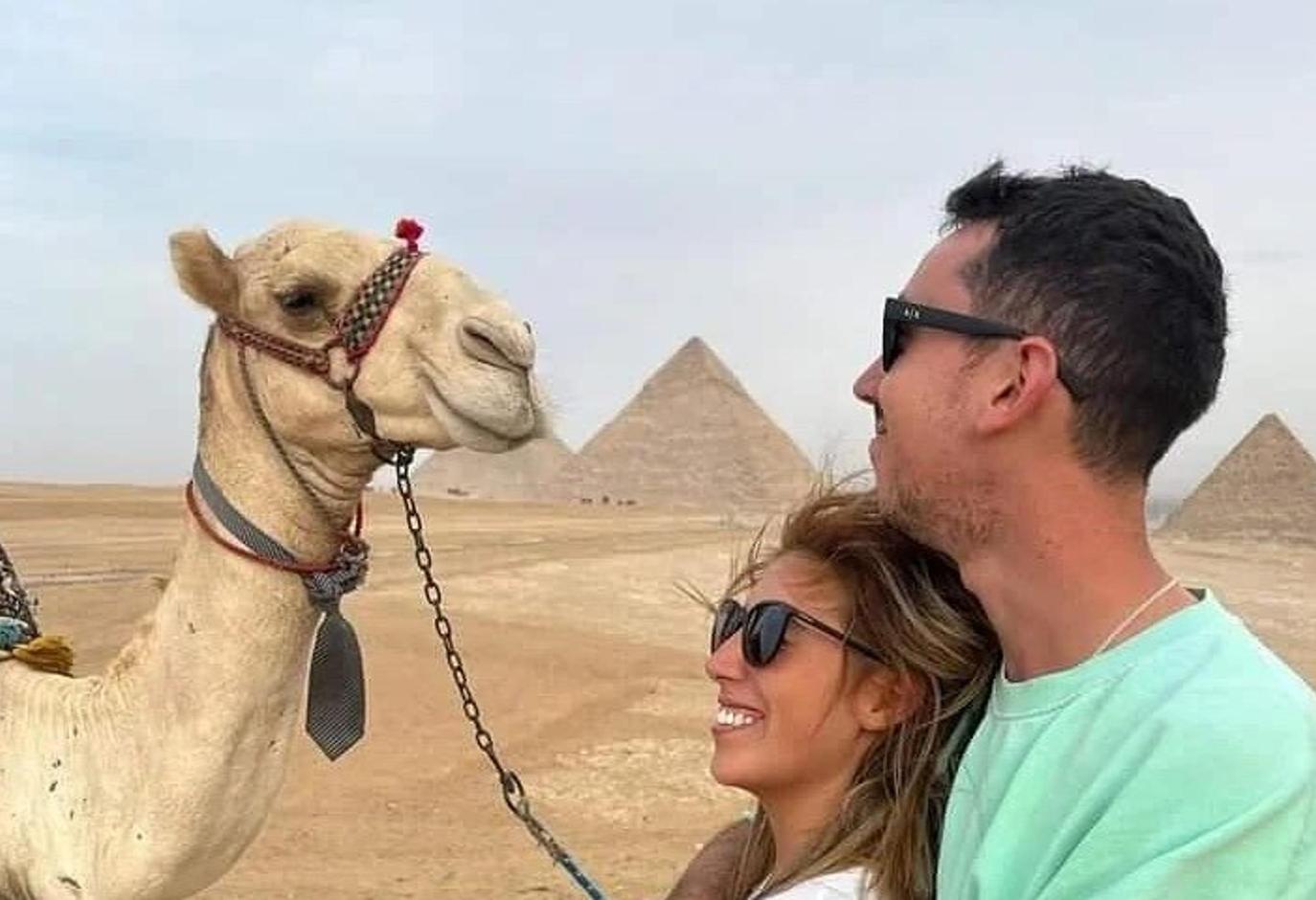 Un hombre y una mujer de pie junto a un camello frente a las pirámides.