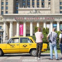 three men standing in front of a yellow car in front of a building