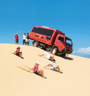 กลุ่มคนนั่งอยู่บนพื้นทรายหน้ารถบรรทุกสีแดง