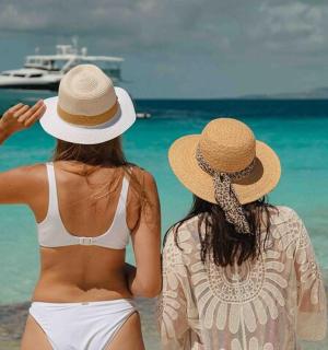 two women sitting on the beach looking at a boat