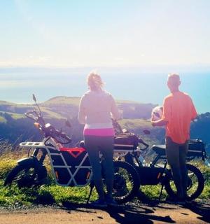 一男一女站在山坡上的一辆摩托车旁。