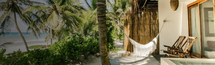 The sun is shining on the verandah of a beach house. On the verandah are a couple of wooden deck chairs, a small swimming pool and a hammock. The hammock is strung between the white wall of the beach house and a palm tree which provides some dappled shade. The beach seems to be accessible from the verandah, just beyond a thicket of palm trees through which the ocean can be seen.