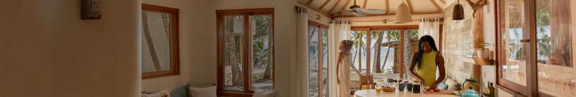 A bright, inviting kitchen with a natural wood and stone design opens up to a tropical beach view through large glass doors. Soft curtains frame the windows, letting in golden sunlight. Two women are inside; one, in a flowing dress, stands near the doors reaching toward a chair, while the other, in a summer top, chops fresh fruit at the kitchen island. The countertop holds a teapot, a pineapple, and other kitchen essentials. Beyond the doors, palm trees and a sandy beach stretch toward the ocean.