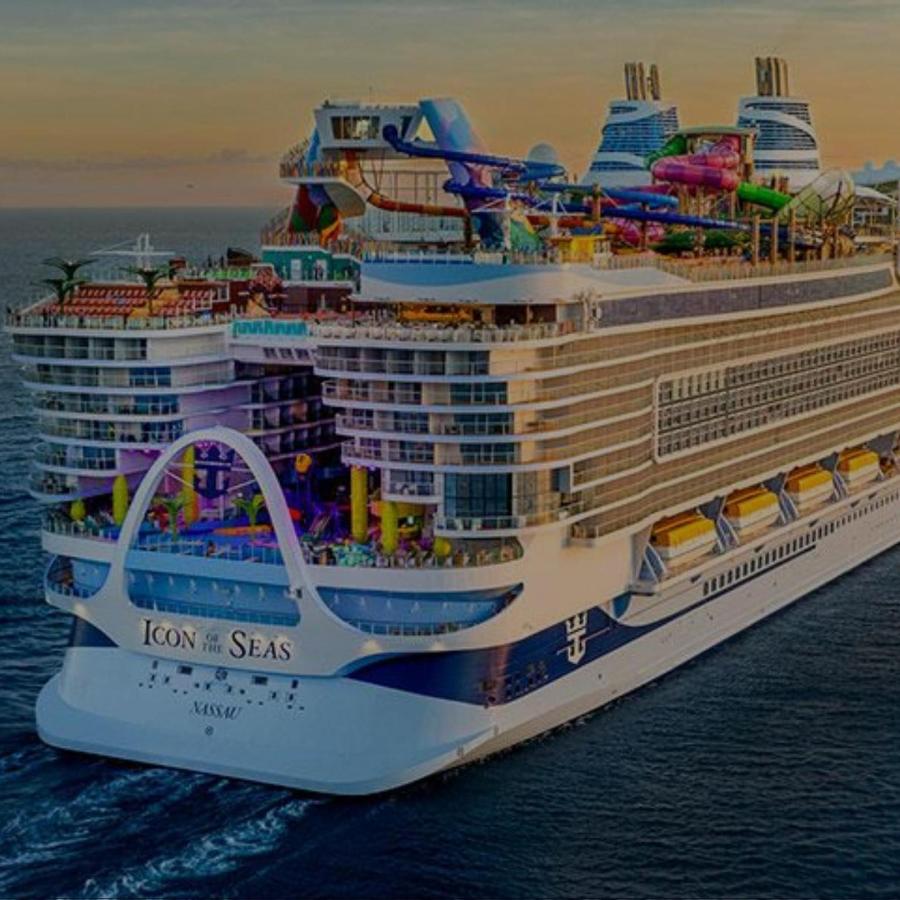 High-angle view of the Royal Caribbean cruise ship Icon of the Seas navigating calm blue ocean waters. Bright multicolored water slides visible on the top deck and rows of passenger balconies lining the side beneath a warm, golden horizon.