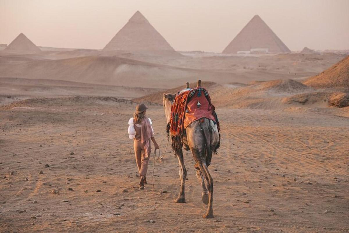eine Frau, die ein Kamel durch die Wüste führt, im Hintergrund sind Pyramiden zu sehen
