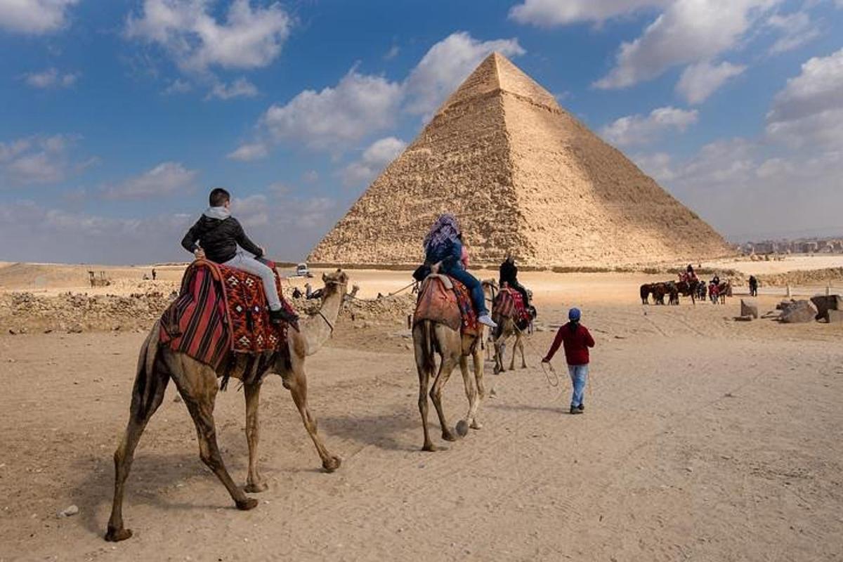 tři lidé jedoucí na velbloudech před pyramidami