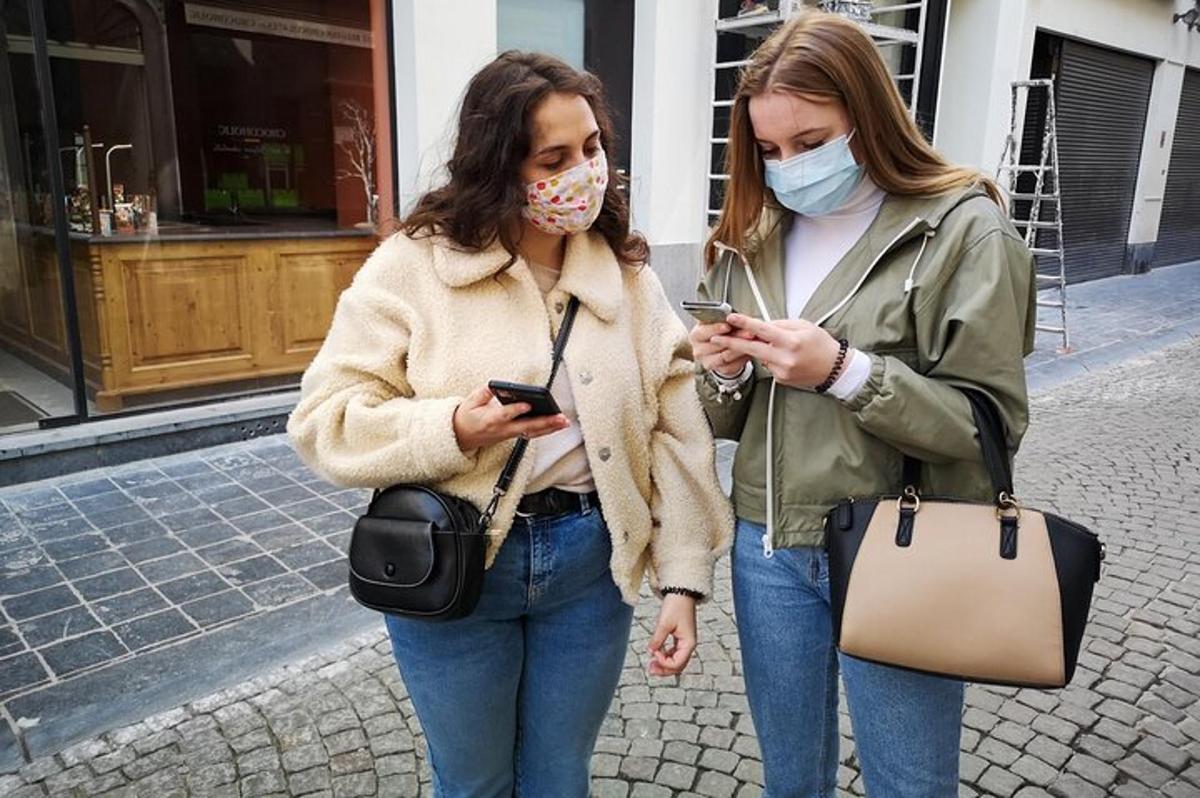 dve ženski z maskami, ki hodita po ulici s svojimi mobilnimi telefoni