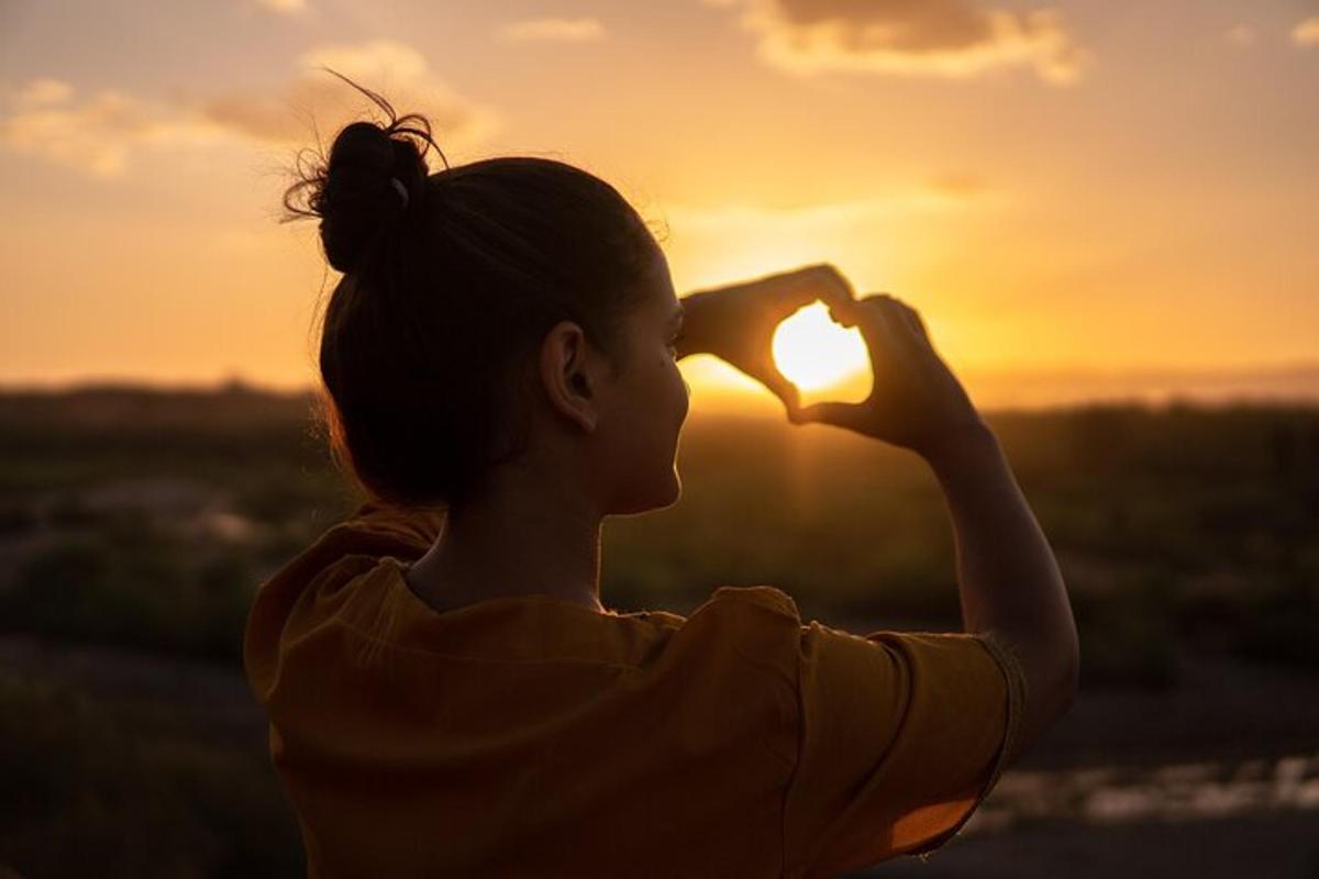 Una mujer haciendo forma de corazón con la puesta de sol de fondo.
