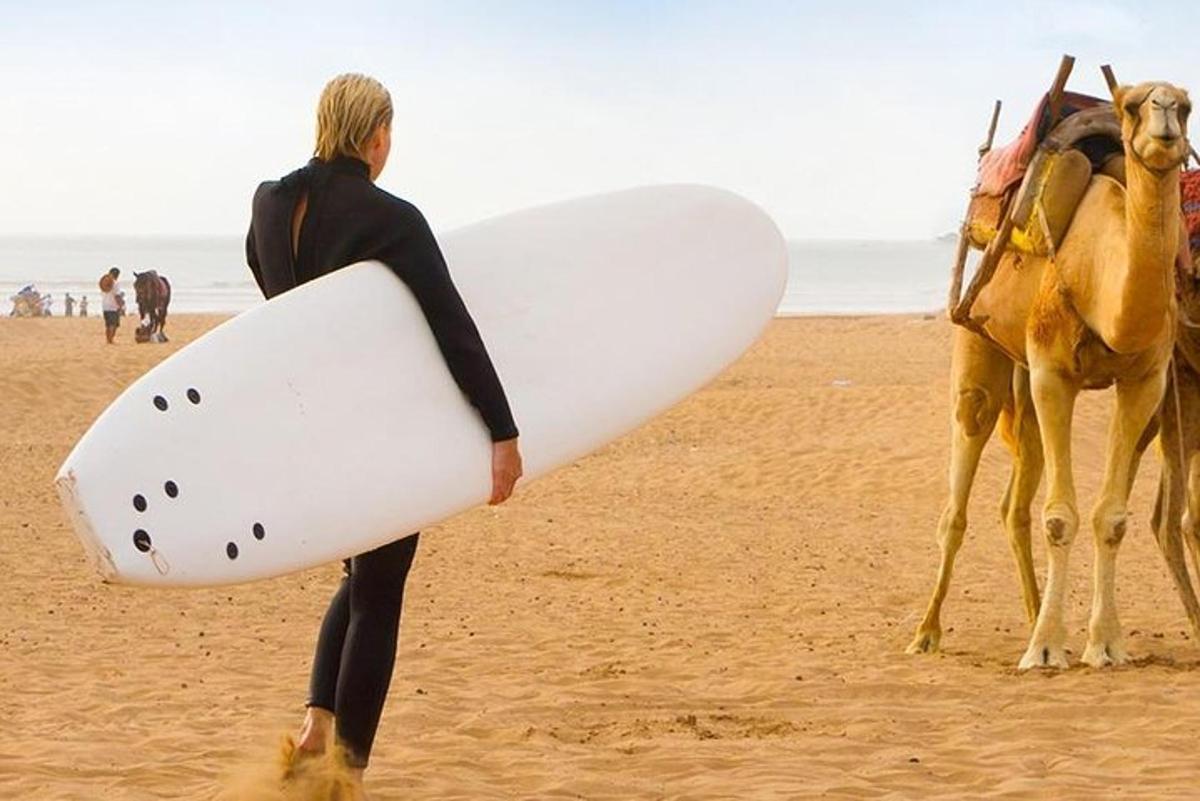 a man holding a surfboard on a beach with a camel