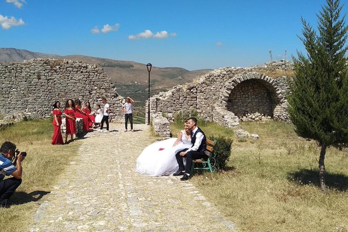 μια νύφη και ένας γαμπρός κάθονται σε ένα παγκάκι δίπλα σε έναν πέτρινο τοίχο
