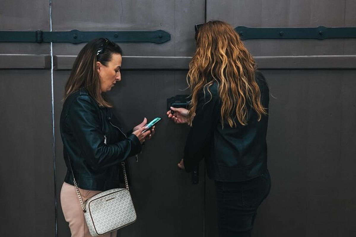 two women standing next to a wall looking at their cell phones