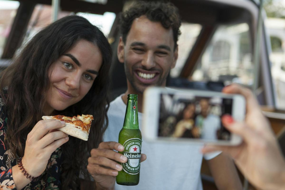 žena fotí muže držícího lahev piva a pizzu