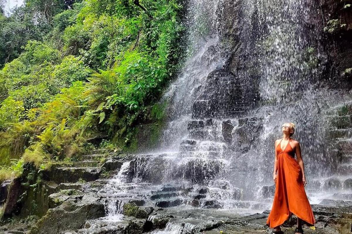 seorang wanita berpakaian oren berdiri di hadapan air terjun