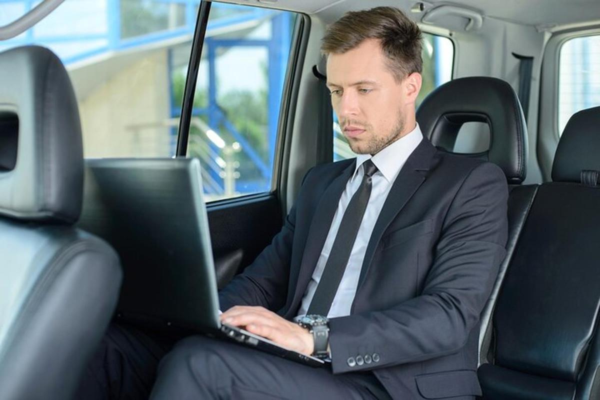 Un hombre con traje y corbata sentado en un automóvil usando una computadora portátil.