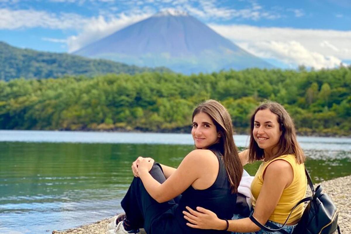 two women sitting next to a lake with a mountain in the background