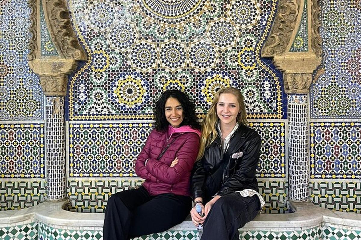 two women sitting on a bench in front of a tile wall