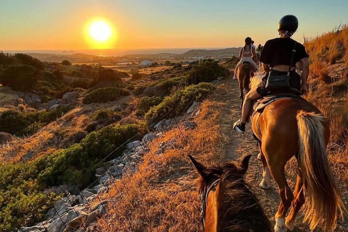 two people riding horses down a hill at sunset