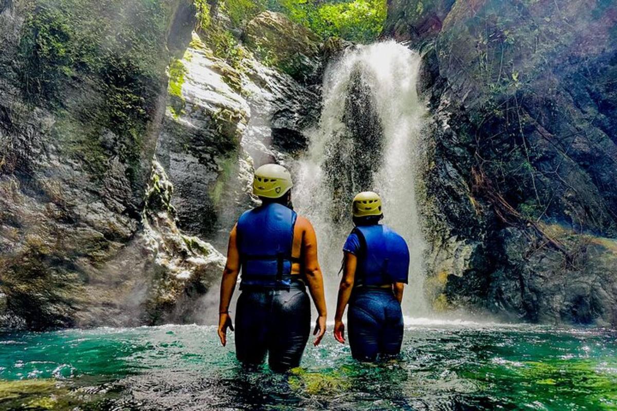 two people walking in the water in front of a waterfall