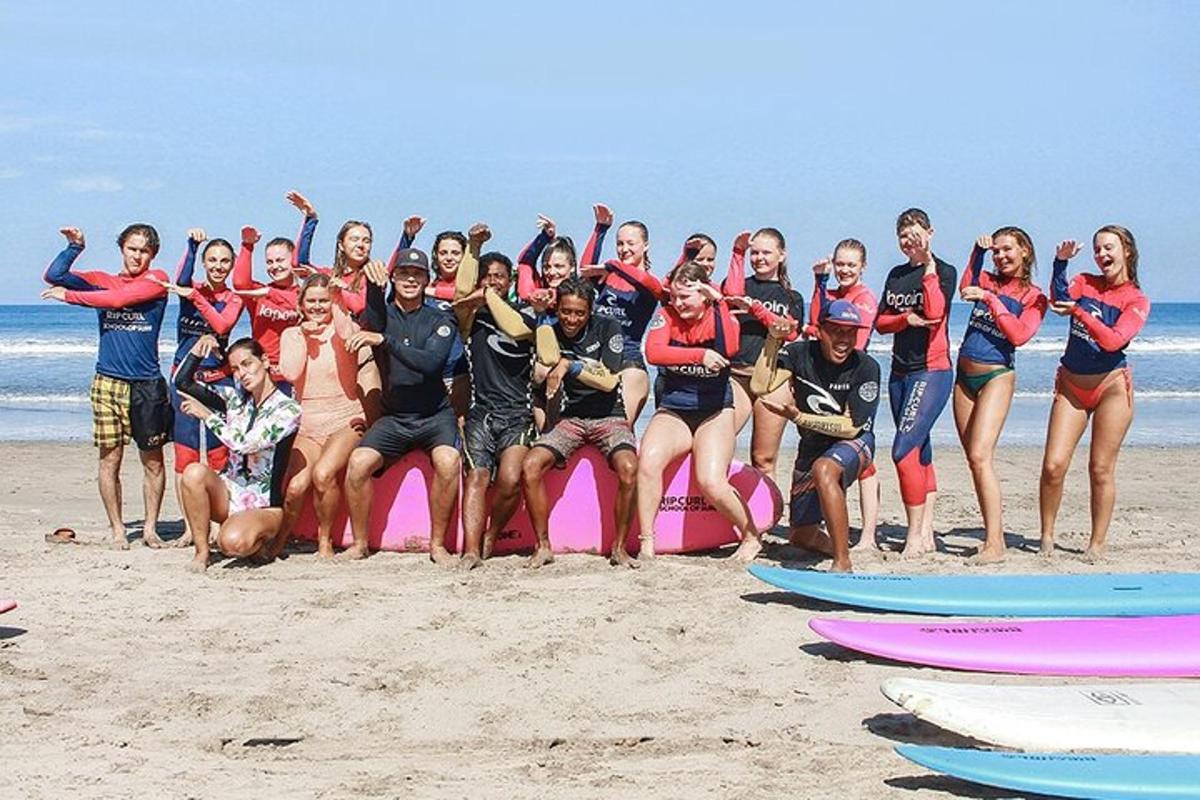 Egy csoport ember pózol egy rózsaszín szörfdeszkán a strandon