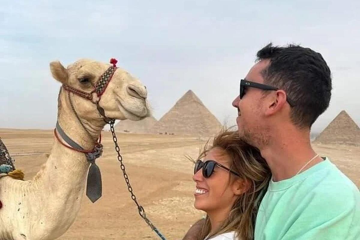 Un hombre y una mujer de pie junto a un camello frente a las pirámides.