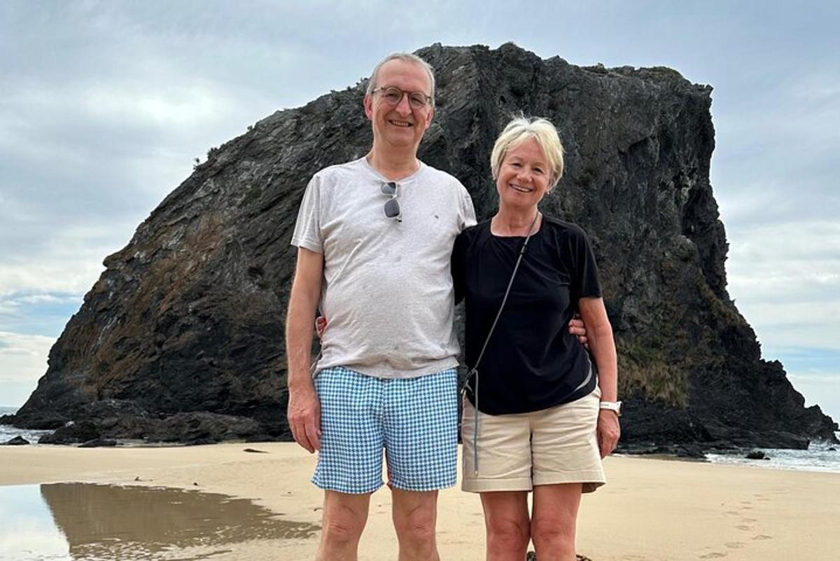 a man and woman standing on the beach near a rock