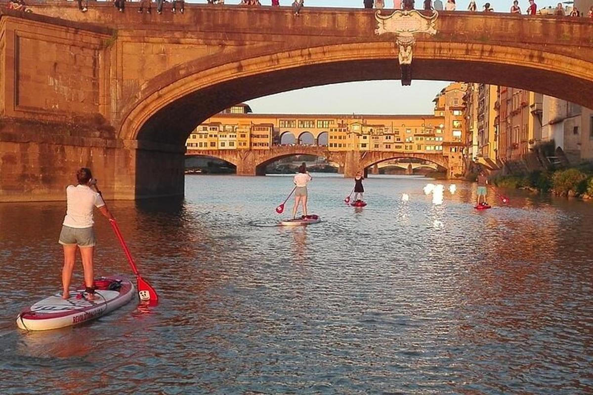 três pessoas em pranchas de remo na água sob uma ponte